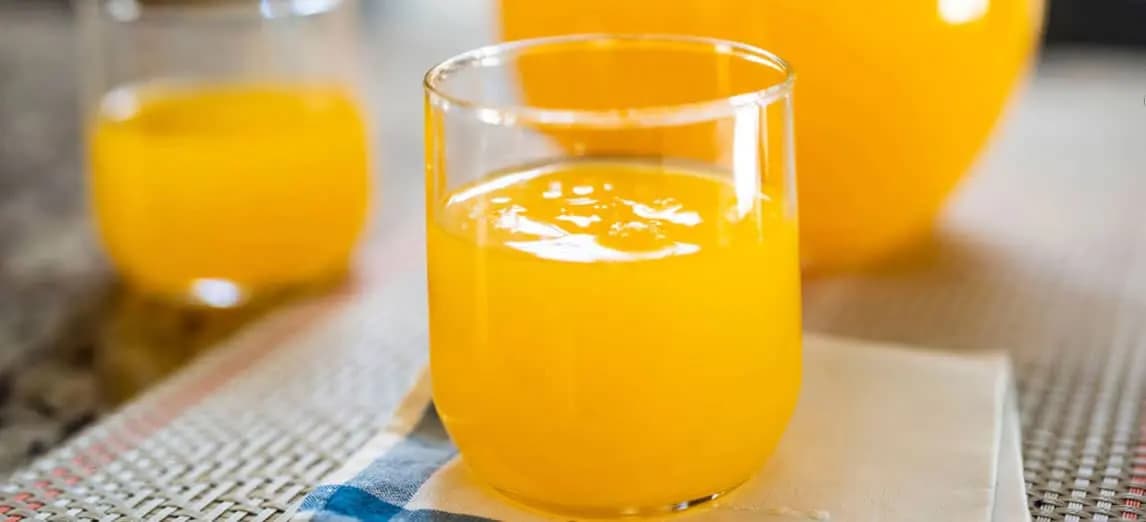 A glass and a smaller cup of bright orange juice, with a pitcher in the background, set on a textured tablecloth