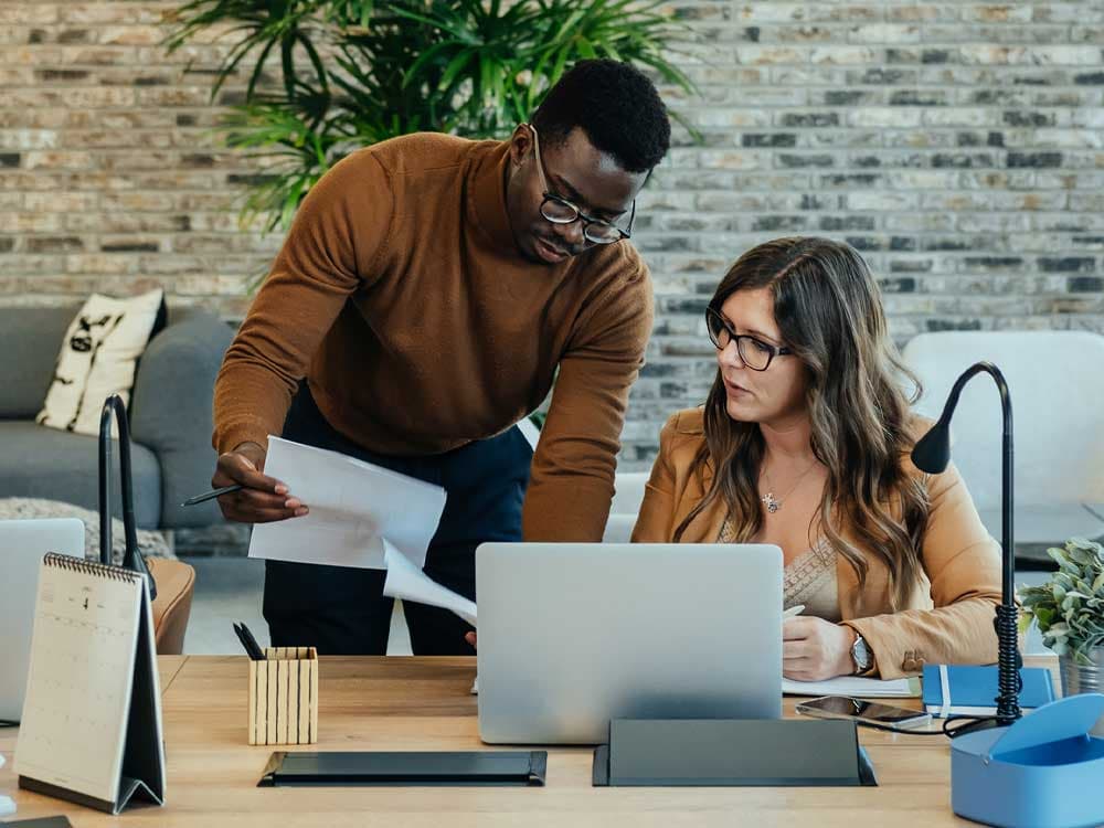 A woman with a smile sitting at a table with a laptop, while a man is positioned in the background. The setting appears to be an indoor office environment.