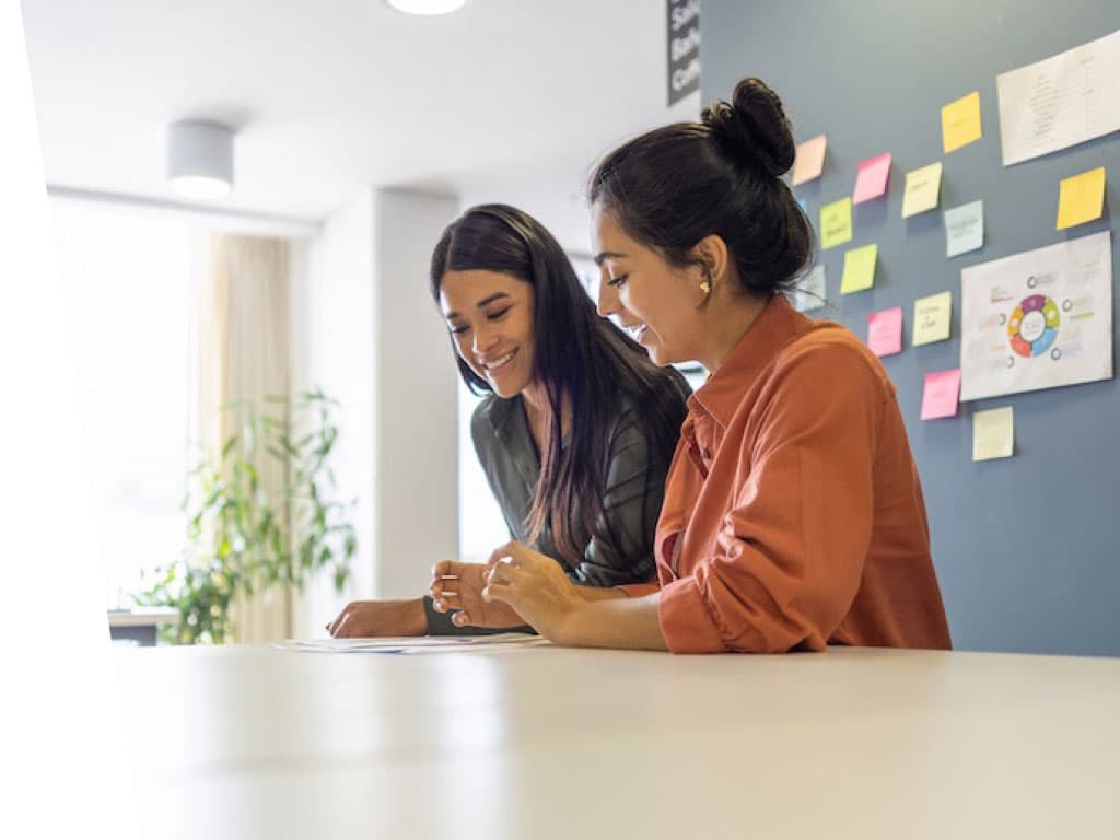 Two women sitting at a table, attentive as they look at a piece of paper in front of them. The setting appears to be an indoor office or classroom with a whiteboard and various office-related elements in the background.