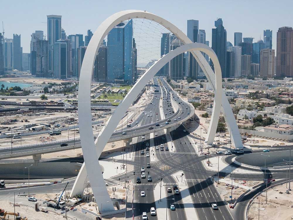 A large arch bridge filled with numerous cars, set against a backdrop of a clear sky and a bustling city skyline featuring skyscrapers and urban buildings.