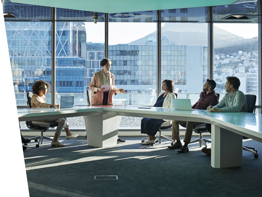 A group of people sitting around a table in an office building, with furniture visible and a window nearby. They are wearing various types of clothing and are engaged in conversation.
