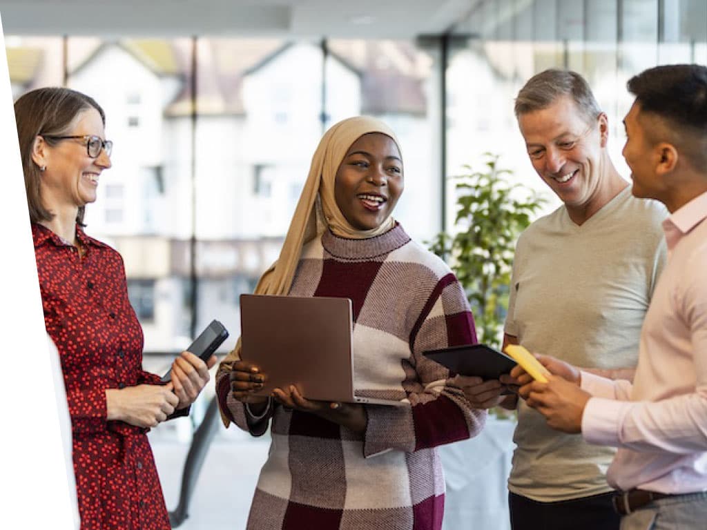 A group of people standing together in a room, smiling and looking engaged. The scene features a mix of men and women, with various clothing styles. The interior has walls that are not specifically described.