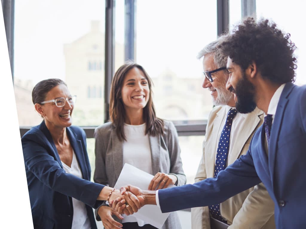 A group of people shaking hands indoors, wearing formal business attire. Some individuals are smiling and engaged in conversation, showcasing collaboration and professionalism.