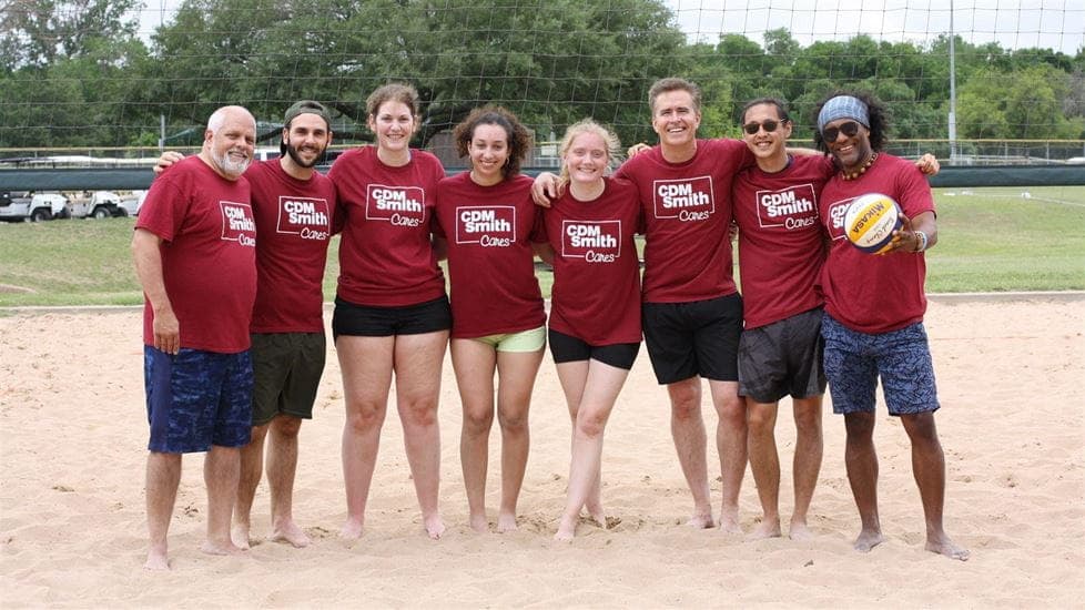 Photograph of a group of eight people standing on a sandy volleyball court, all wearing matching maroon shirts with white text reading "CDM Smith Classic." One person on the far right is holding a volleyball, and the background features green trees and a volleyball net.