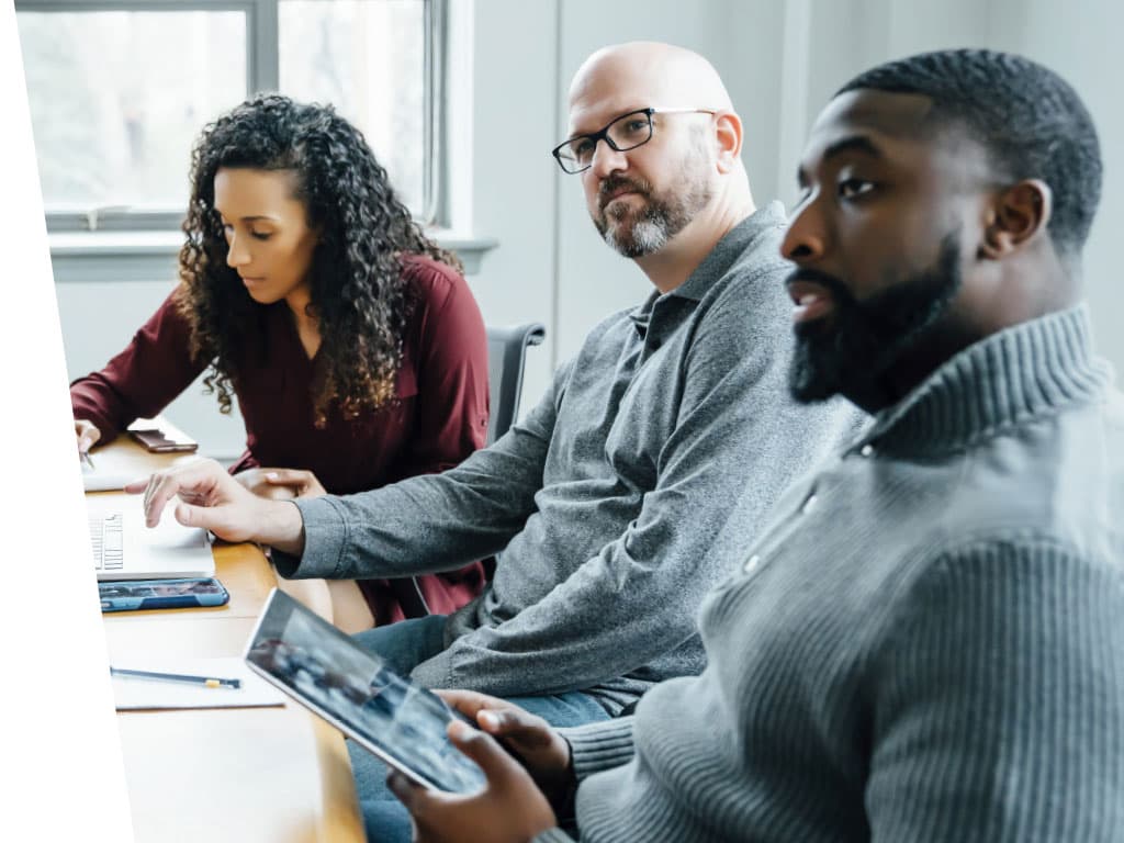 A group of people sitting around a table, attentively looking at a tablet. They exhibit expressions of interest and engagement, with warm lighting and a casual indoor setting.