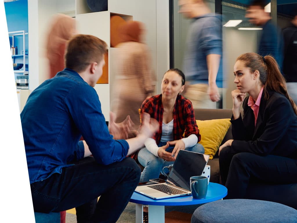 A group of people seated in a circle indoors, engaging with one another. The setting features a wall in the background, and both men and women are present, showcasing a variety of clothing styles and human expressions.