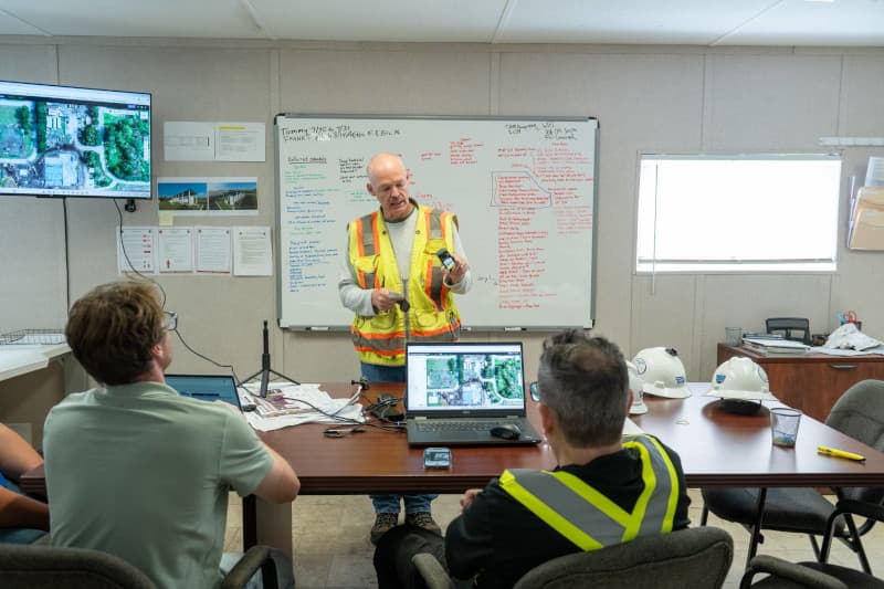 Photograph of two people standing indoors, engaged in discussion while holding a laptop. The setting appears to be a modern office or workspace with blurred background elements suggesting a professional environment.