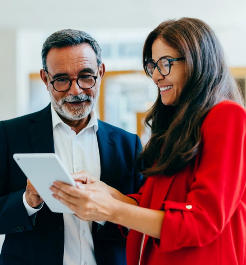 Photograph of two business professionals engaged in a discussion while looking at a tablet. The man wears a dark suit and white shirt, and the woman wears a bright red blazer, indicating a collaborative work environment.