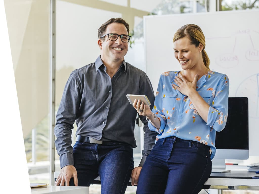 A man and woman are smiling while looking at a phone indoors. The man is wearing a shirt and the woman is wearing glasses and jeans, both standing in front of a wall.