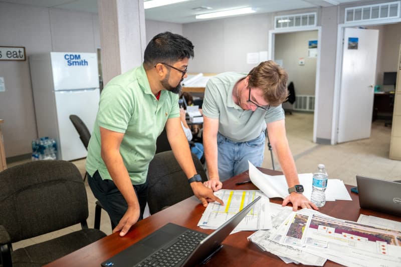 Photograph of two people collaborating in an office setting, reviewing printed documents and plans spread across a table alongside a laptop and water bottle.