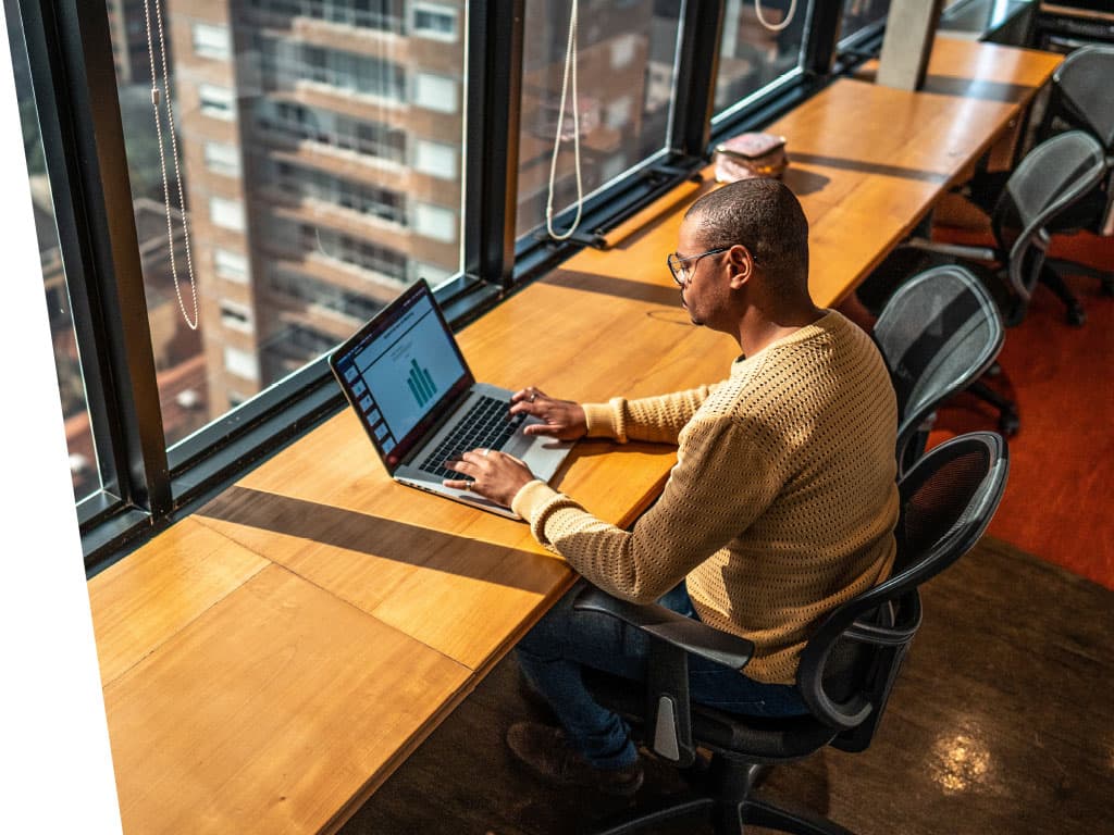 A man sitting at a wooden table in an office building, using a laptop. The room has a window and features various pieces of furniture like a chair and desk.