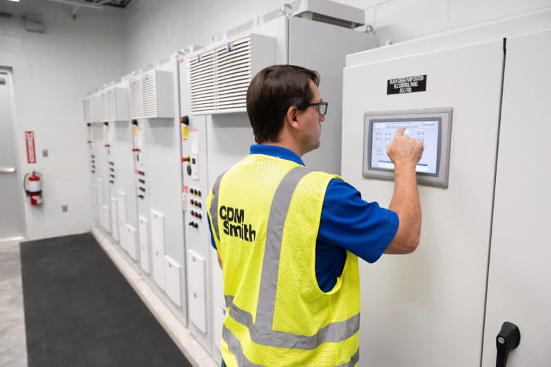 Photograph of a worker wearing a yellow safety vest labeled "CDM Smith" interacting with a touchscreen control panel on a large industrial electrical cabinet.