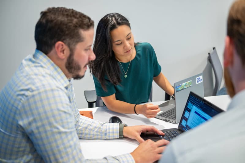 Photograph of three people collaborating around a table with laptops, likely in a meeting or work session.