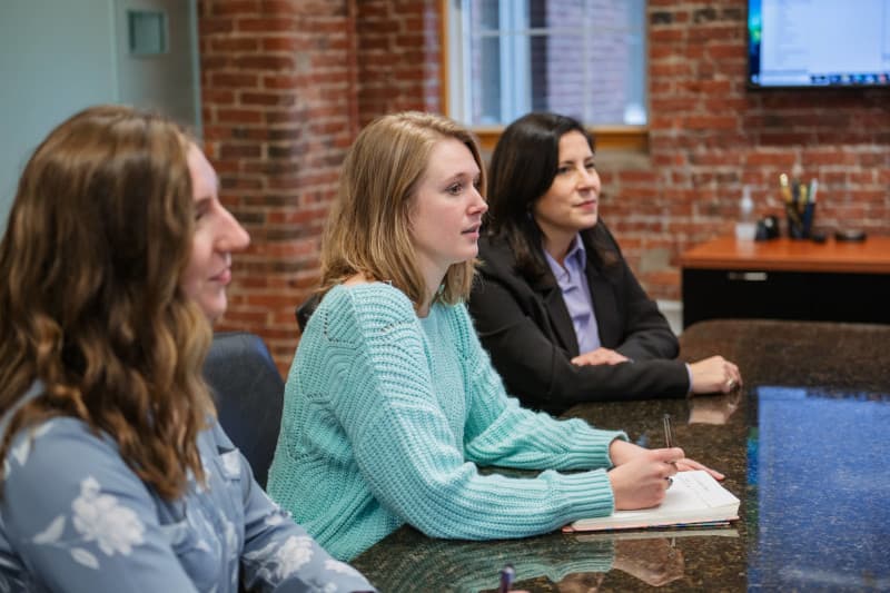 Photograph of three women seated at a conference table in a modern office with exposed brick walls, engaged in a meeting or discussion.