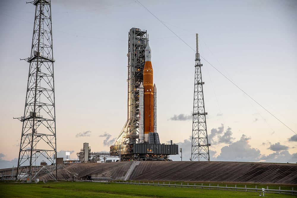 A large orange rocket stands prominently on a grassy field under a clear blue sky.