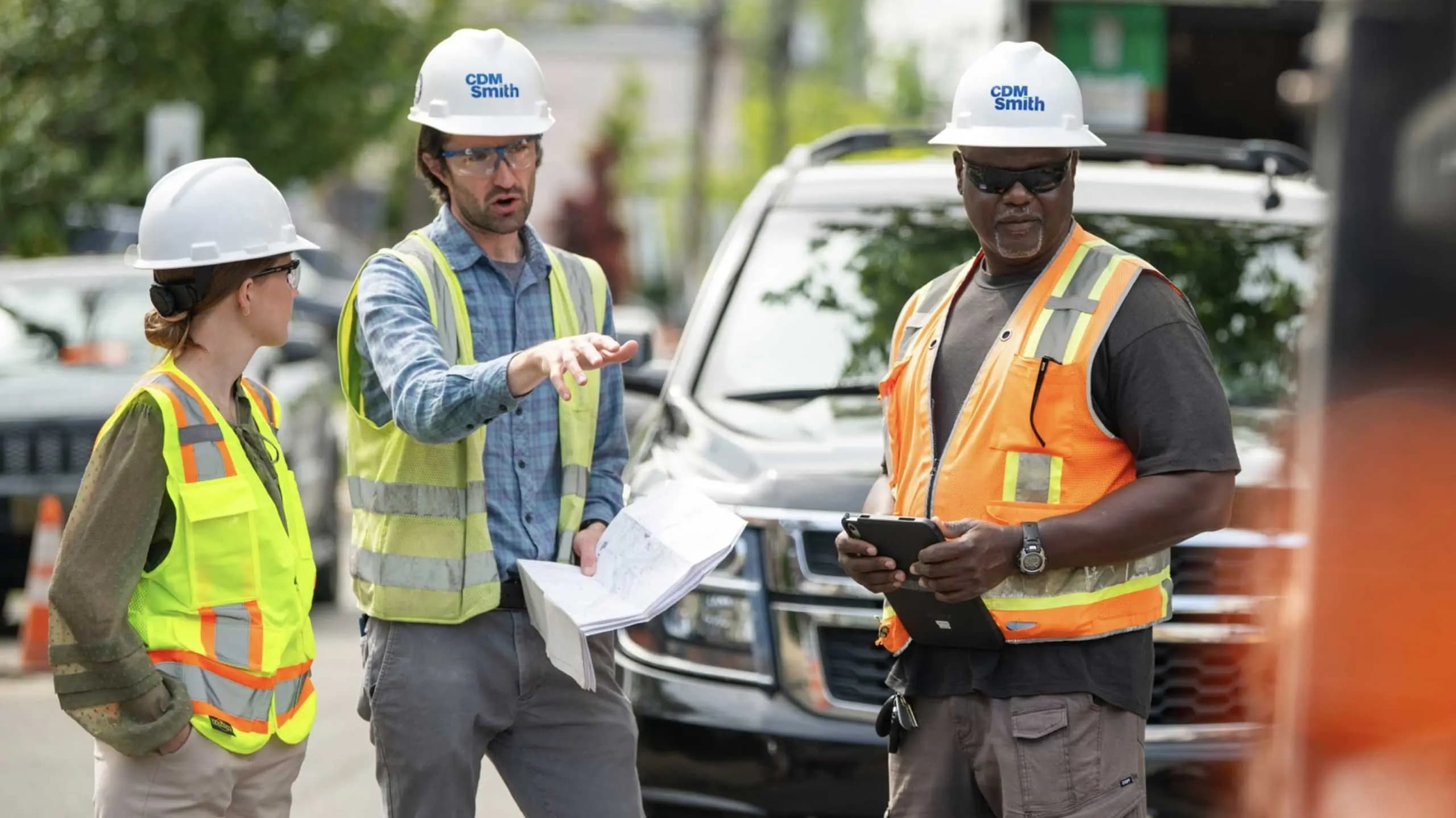 Three construction workers in hard hats standing in front of a parked car on a construction site.