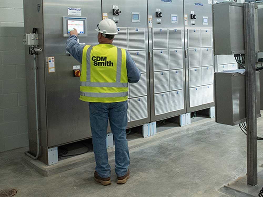 A man wearing a safety vest and white helmet stands indoors, representing a blue-collar worker or engineer in workwear.