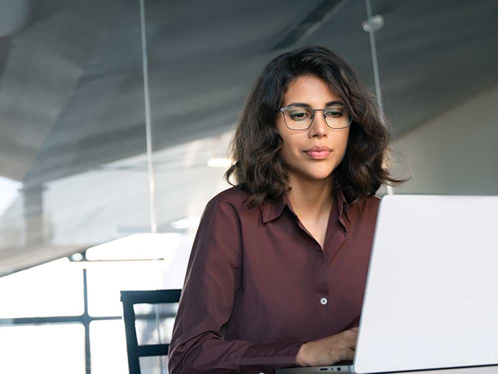 A woman sitting at a desk with a laptop, wearing casual clothing, indoors. The background includes a wall.