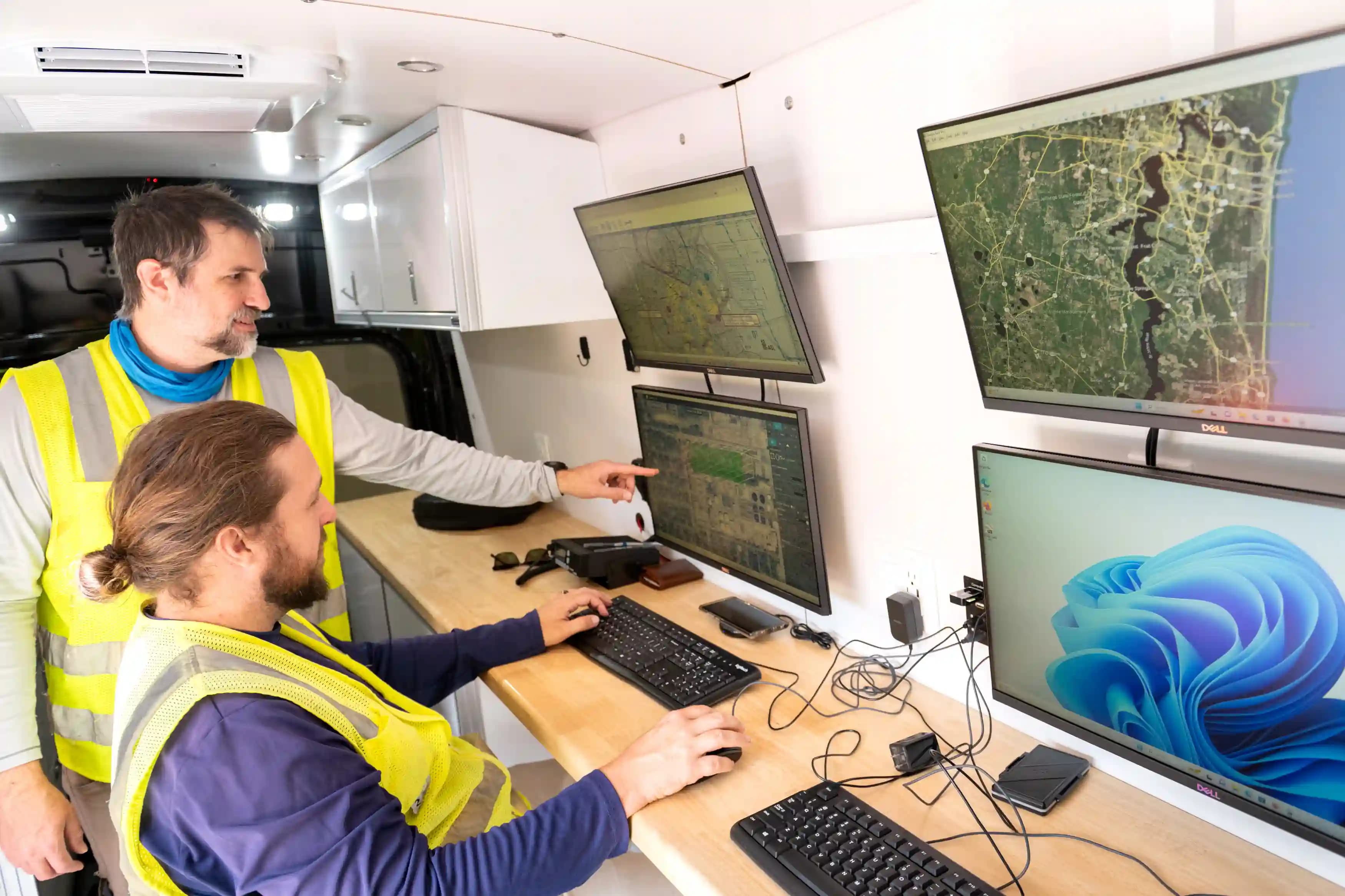Photograph of two people wearing high-visibility vests working inside a mobile command center with multiple monitors displaying maps and a computer desktop screen. The setup includes a keyboard, mouse, and various cables, indicating a focus on navigation, monitoring, or coordination tasks.