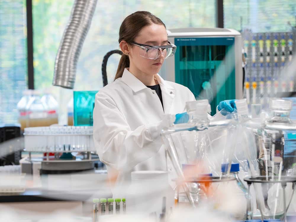 A woman in a white coat and gloves stands in a laboratory, surrounded by various scientific instruments and equipment, engaged in research.