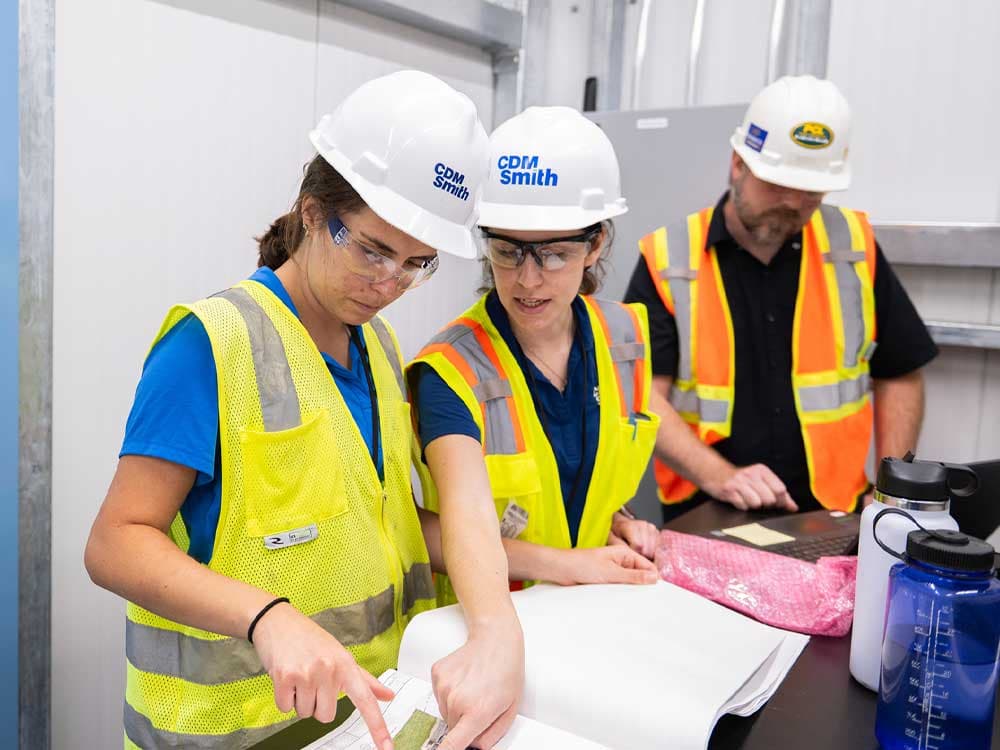 A group of people indoors wearing safety vests and helmets, likely in a construction or engineering setting.
