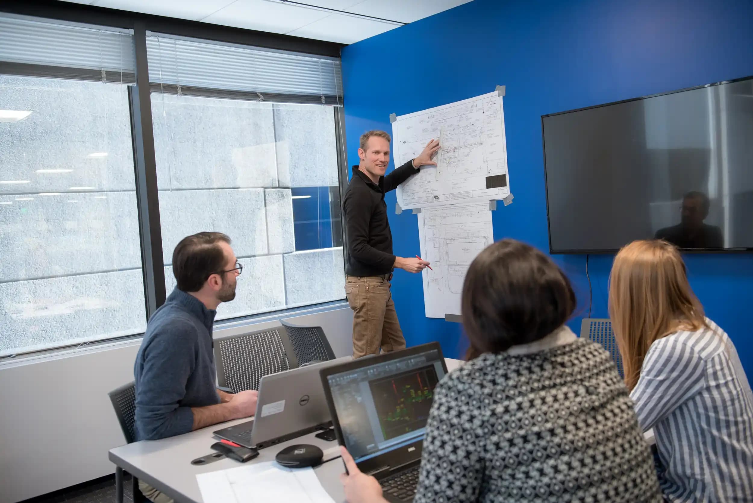 Photograph of a business meeting in a modern office with four people, one standing and pointing at large sheets of paper with diagrams or notes taped to a blue wall. Three seated participants are engaged, with two using laptops, indicating a collaborative discussion or presentation focused on project planning or strategy.