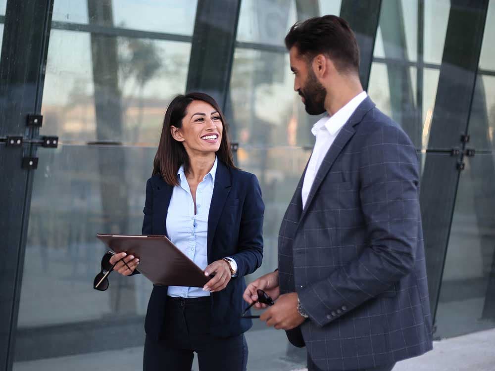 A man and woman standing next to each other outdoors. The man is wearing a blazer and a dress shirt, smiling confidently. The woman is dressed in formal attire, also smiling as they pose together.