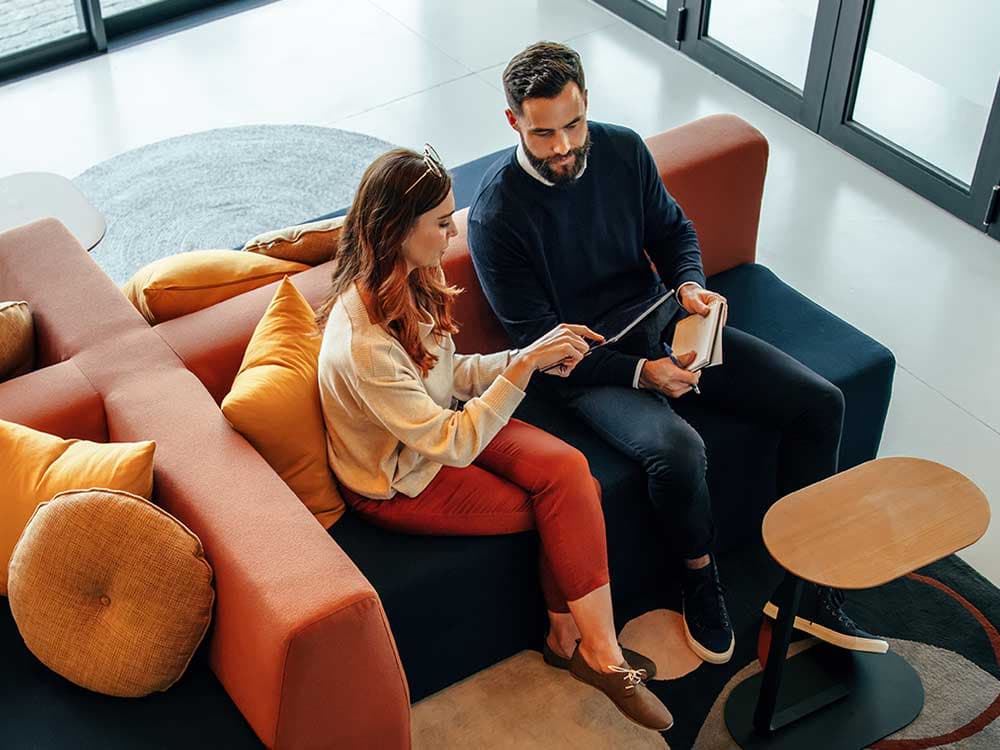 A man wearing a vest is pointing at a computer screen while seated at a desk with a personal computer.