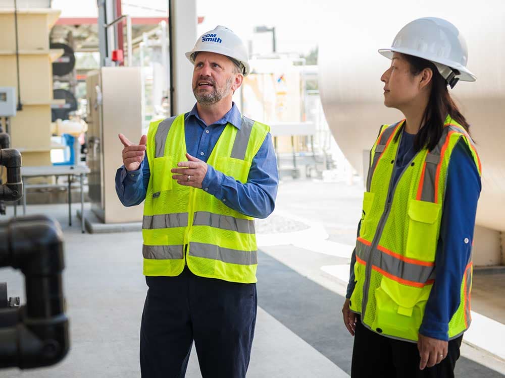 A man and woman wearing safety vests and helmets standing indoors.