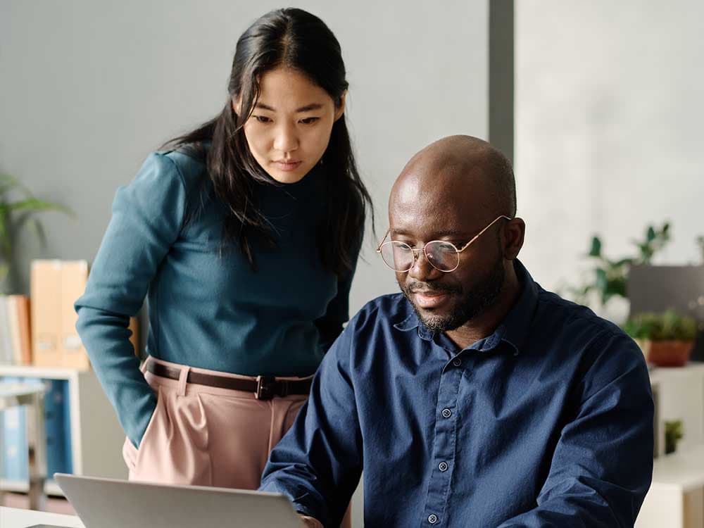 A man and a woman are standing indoors, looking at a tablet together. The man is wearing a dress shirt and is smiling, while the woman also appears to be engaged with the screen. A wall is visible in the background.