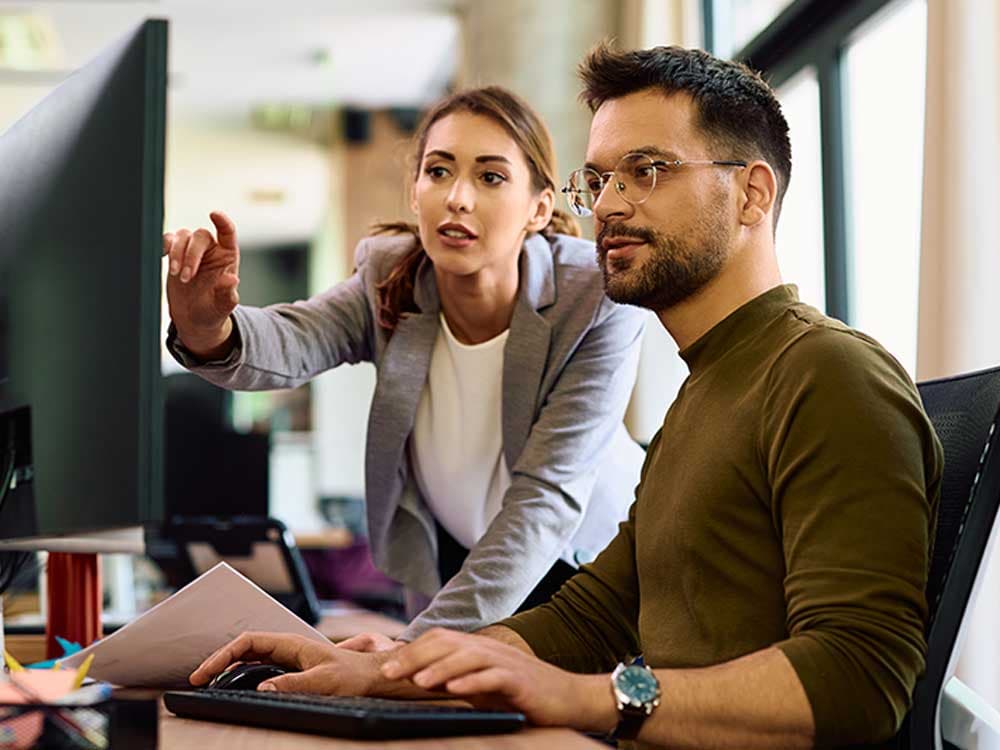 A woman in an indoor office setting is pointing at a laptop screen while sitting at a desk.