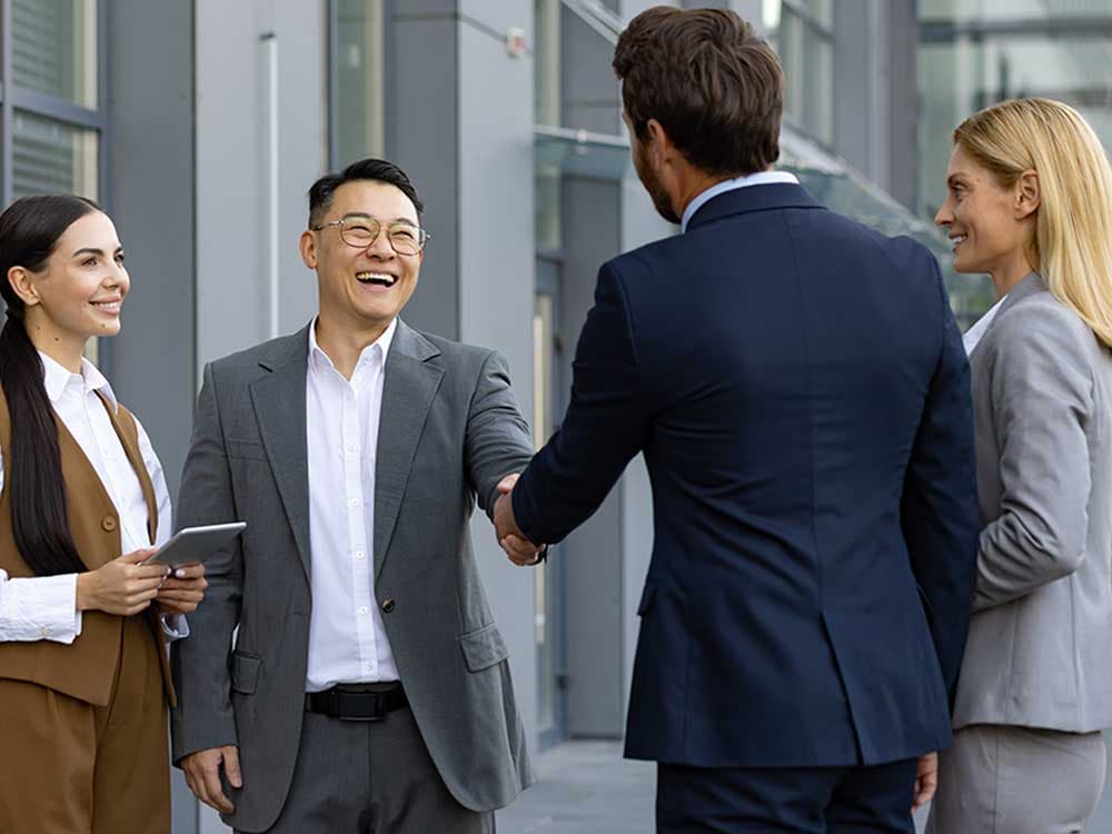 A man in a suit shakes hands with another man, both smiling, in an outdoor setting. They are dressed in formal wear and appear to be engaged in a professional collaboration.