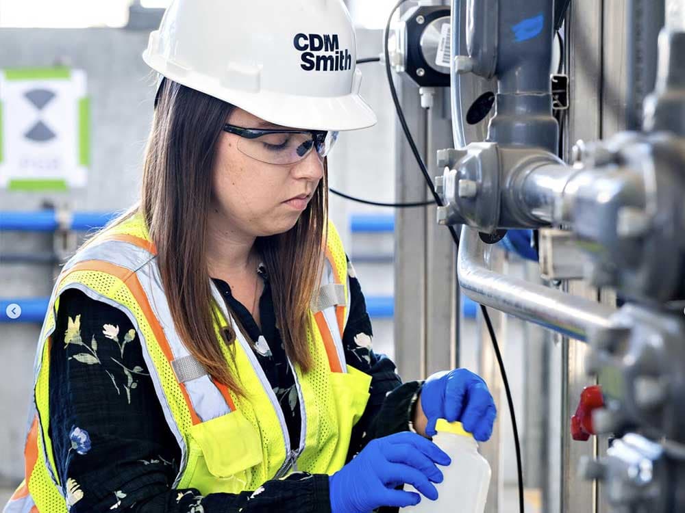 A woman wearing a safety vest and gloves stands outdoors, equipped with a helmet and personal protective equipment, representing a technician or engineer in blue-collar work.