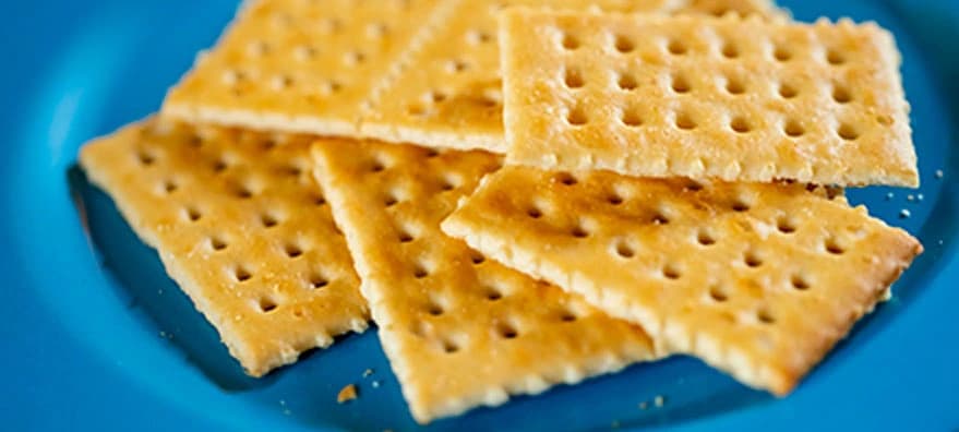 A stack of golden-brown crackers on a bright blue plate, arranged neatly with a textured surface and perforations visible