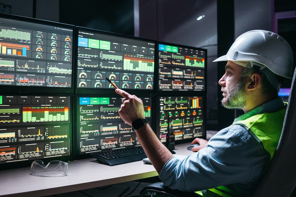 A man wearing a hard hat studies several screens, likely analyzing information and monitoring a project.