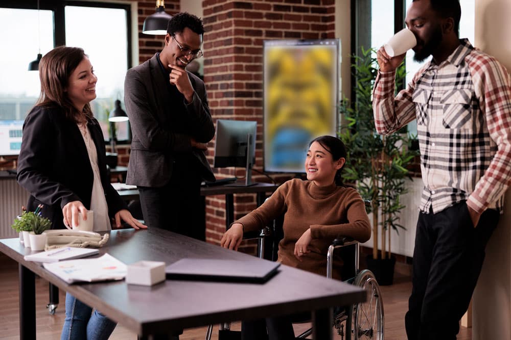 A diverse group of people gathers around a table, with a woman in a wheelchair actively participating in the discussion.