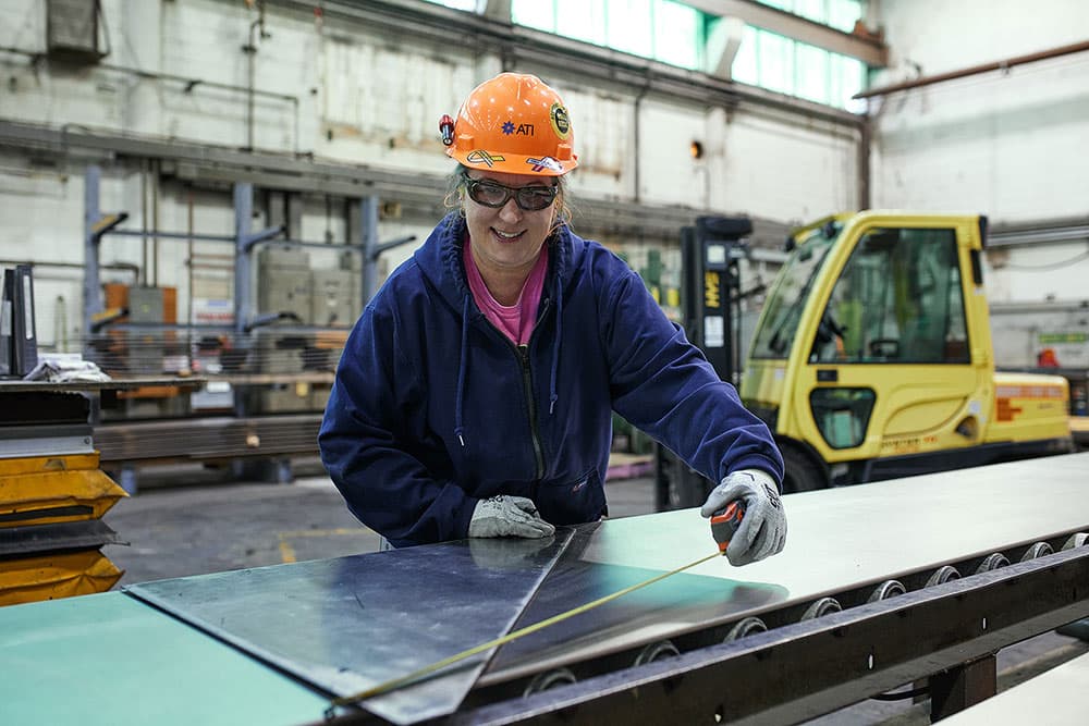 A woman engaged in work at a metal table inside a factory, surrounded by industrial equipment.