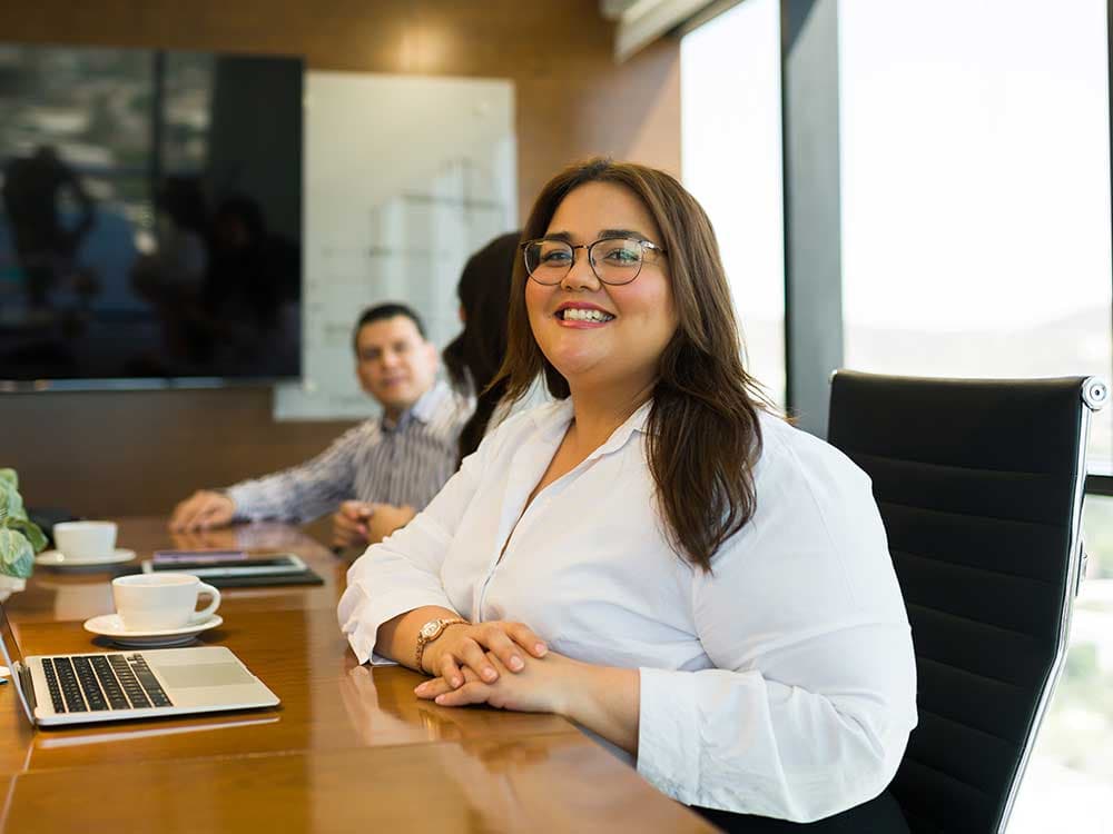 A woman with a smile sitting at a table with a laptop, while a man is positioned in the background. The setting appears to be an indoor office environment.