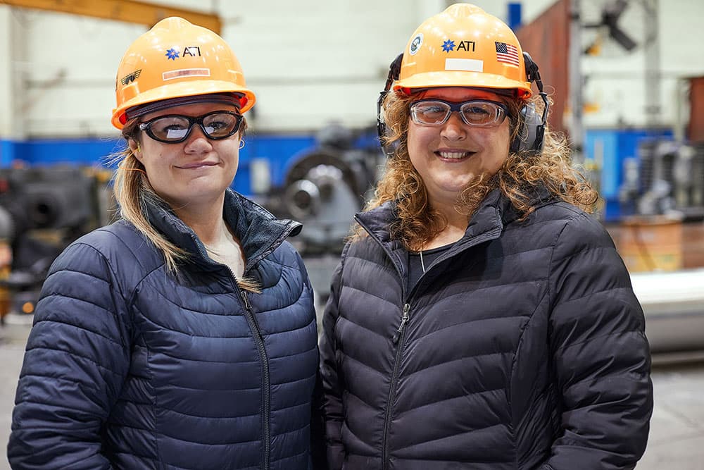 Two women wearing hard hats and glasses pose in front of a machine.