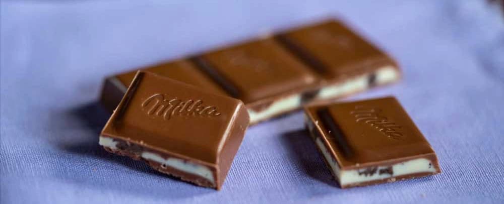 Nærbillede af afbrækket Milka-chokolade på en lavendelblå dug, der viser blød og lækker chokolade med creme indeni
