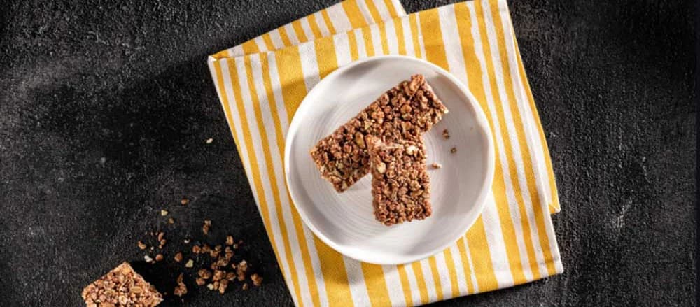 Granola-stænger på en hvid tallerken, der står på en gul- og hvidstribet serviet med krummer rundt omkring på en sort baggrund