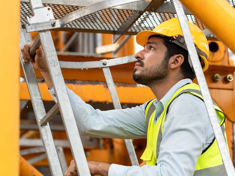 A man wearing a hard hat and high-visibility vest climbs a ladder outdoors.